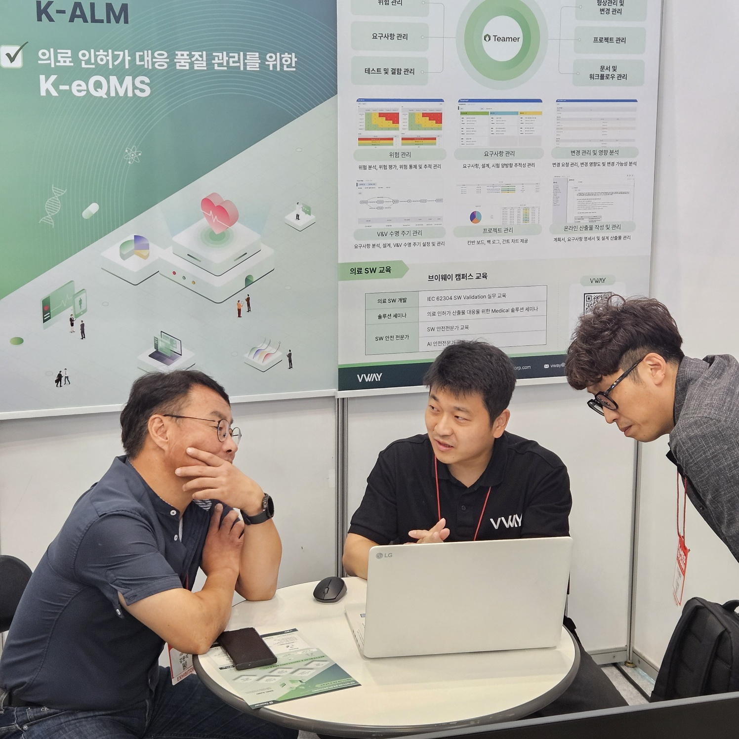 KIMES Busan 2024 브이웨이 부스 상담 장면 — 뒤편 패널에 Teamer 의 위험 관리, 요구사항 관리, 변경 관리 및 영향 분석, V&V 수명 주기 관리, 프로젝트 관리, 온라인 산출물 작성 및 관리 기능과, 브이웨이 캠퍼스 의료 SW 교육 과정(IEC 62304 SW Validation 실무 교육, Medical 솔루션 세미나, SW·AI 안전전문가 교육) 안내