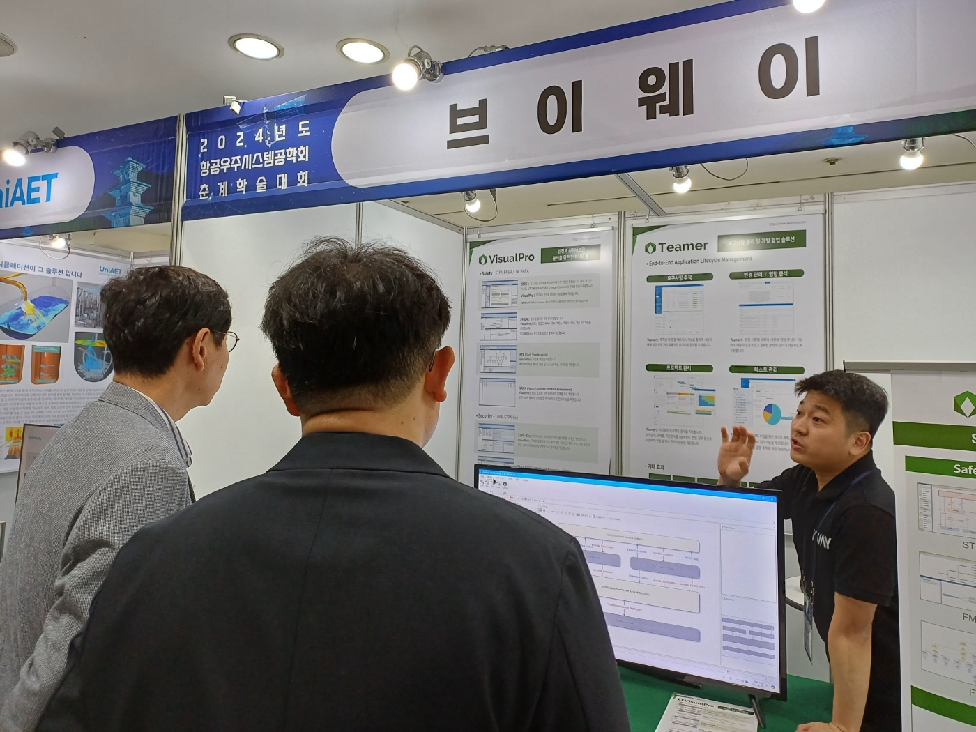 2024년도 항공우주시스템공학회 춘계학술대회 브이웨이 부스 — VisualPro(STPA·FMEA·FTA·HARA·TARA·STPA-Sec)와 Teamer(End-to-End ALM) 패널 앞에서 방문객에게 컨트롤 스트럭처 화면을 설명하는 모습
