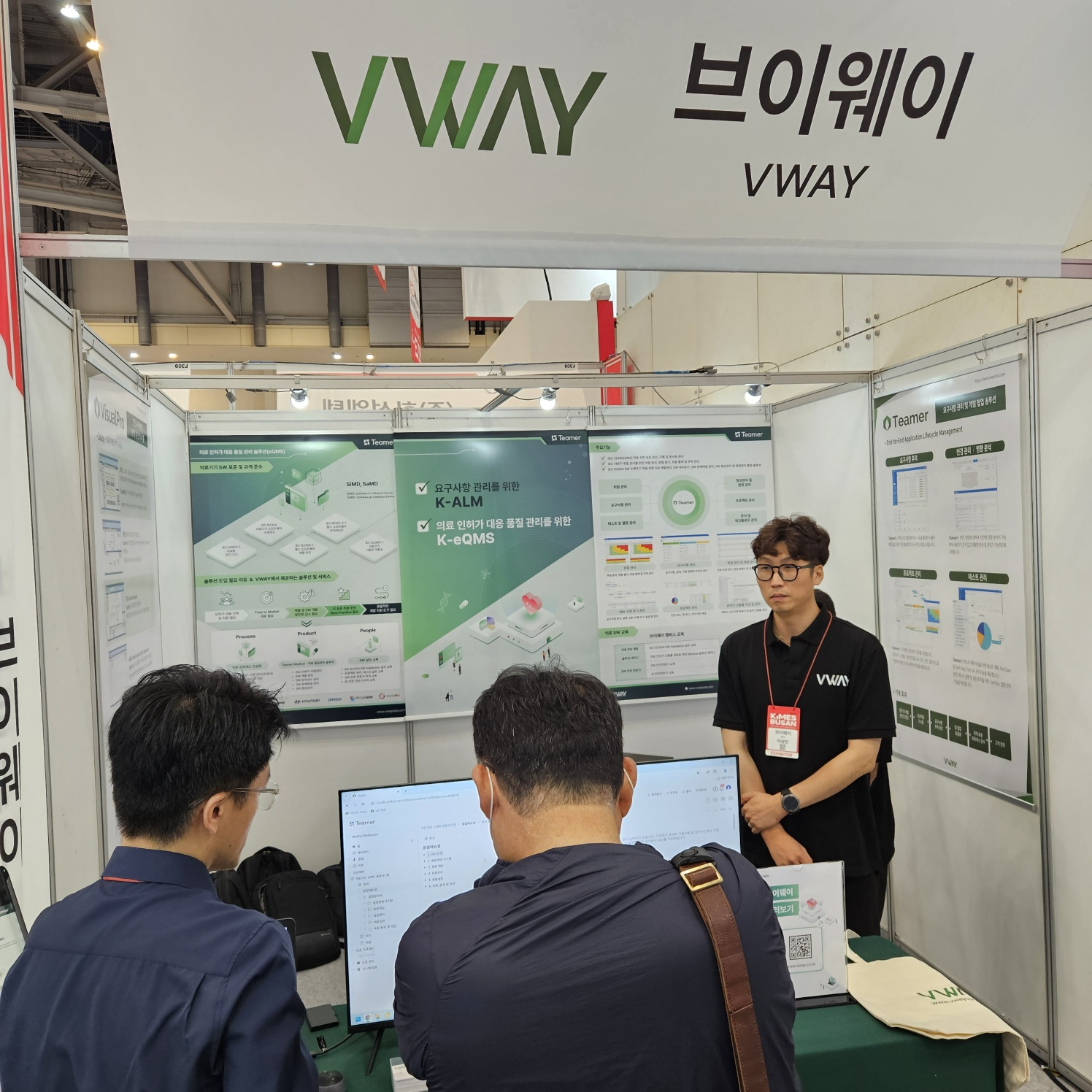KIMES Busan 2024 브이웨이 부스 — 의료 인허가 대응 품질관리 솔루션(eQMS) 패널, 요구사항 관리를 위한 K-ALM 과 의료 인허가 대응 품질 관리를 위한 K-eQMS 안내, Teamer 요구사항 추적·변경 관리·프로젝트 관리·테스트 관리 패널. 방문객에게 Teamer 화면을 설명하는 모습