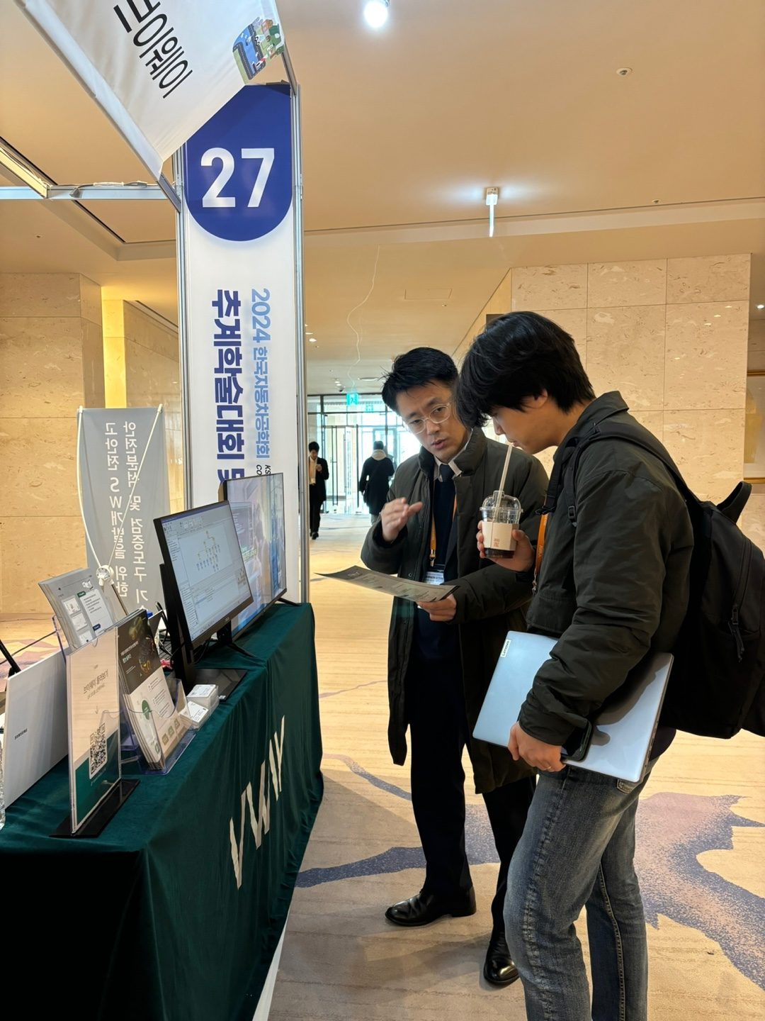 2024 한국자동차공학회 추계학술대회 브이웨이 부스(27번) — 방문객에게 브로슈어로 안전분석 솔루션을 설명하는 모습, 모니터에 VisualPro 분석 화면