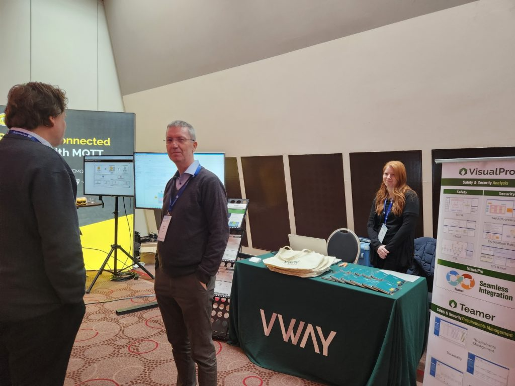 Automotive Cyber Security Europe 2023 브이웨이 부스 — VisualPro 안전·보안 분석과 Teamer 요구사항 관리 배너, 모니터에 분석 화면. 방문객과 대화하는 직원들