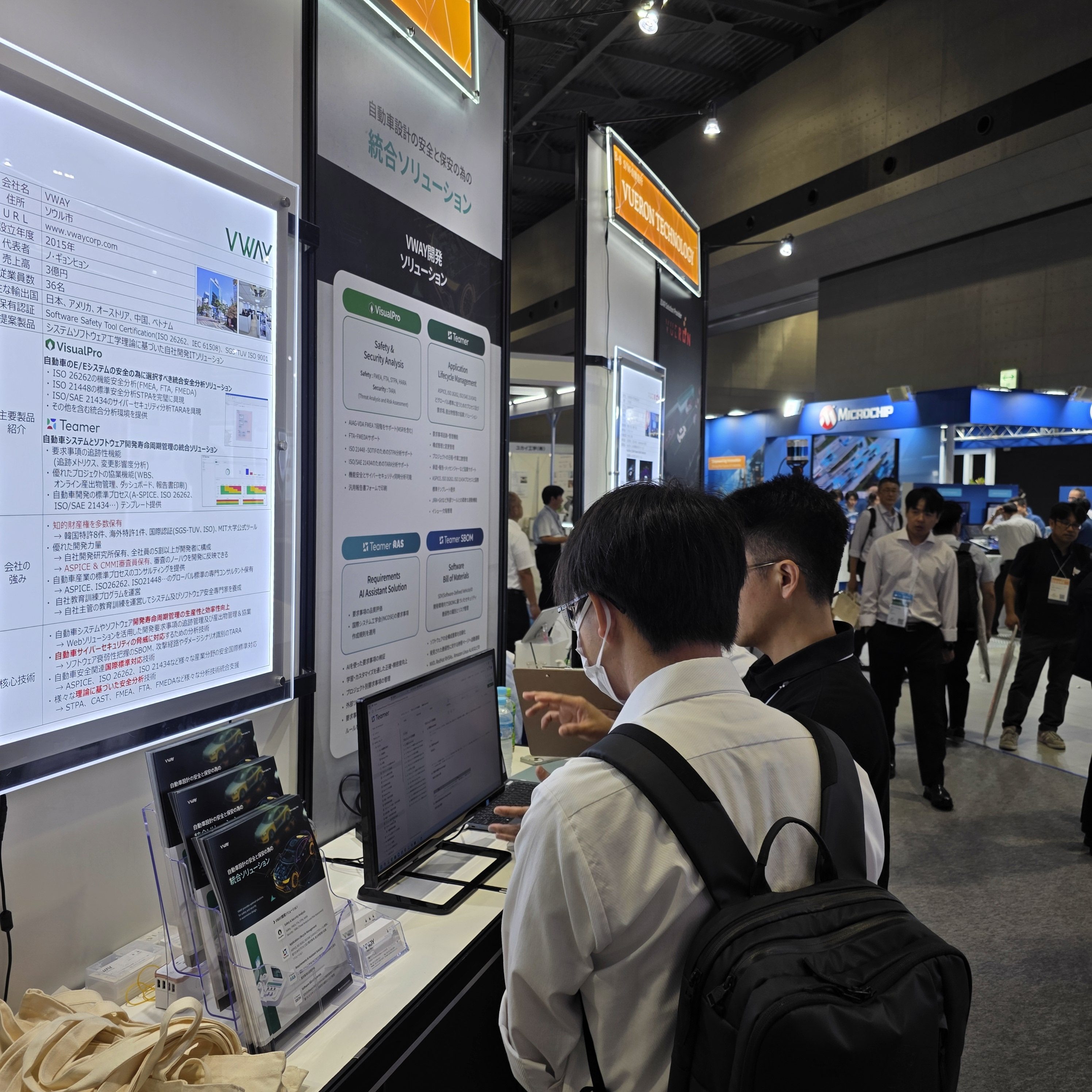 Automotive Engineering Exposition 2025 Nagoya 브이웨이 부스에서 일본 방문객이 Teamer 화면을 살펴보는 모습 — 옆 패널에 회사 개요(설립 2015년, 대표 노경현, 매출 3억엔, 임직원 36명, 주요 수출국 일본·미국·오스트리아·중국·베트남, 보유 인증 Software Safety Tool Certification(ISO 26262·IEC 61508)과 SGS-TUV ISO 9001)