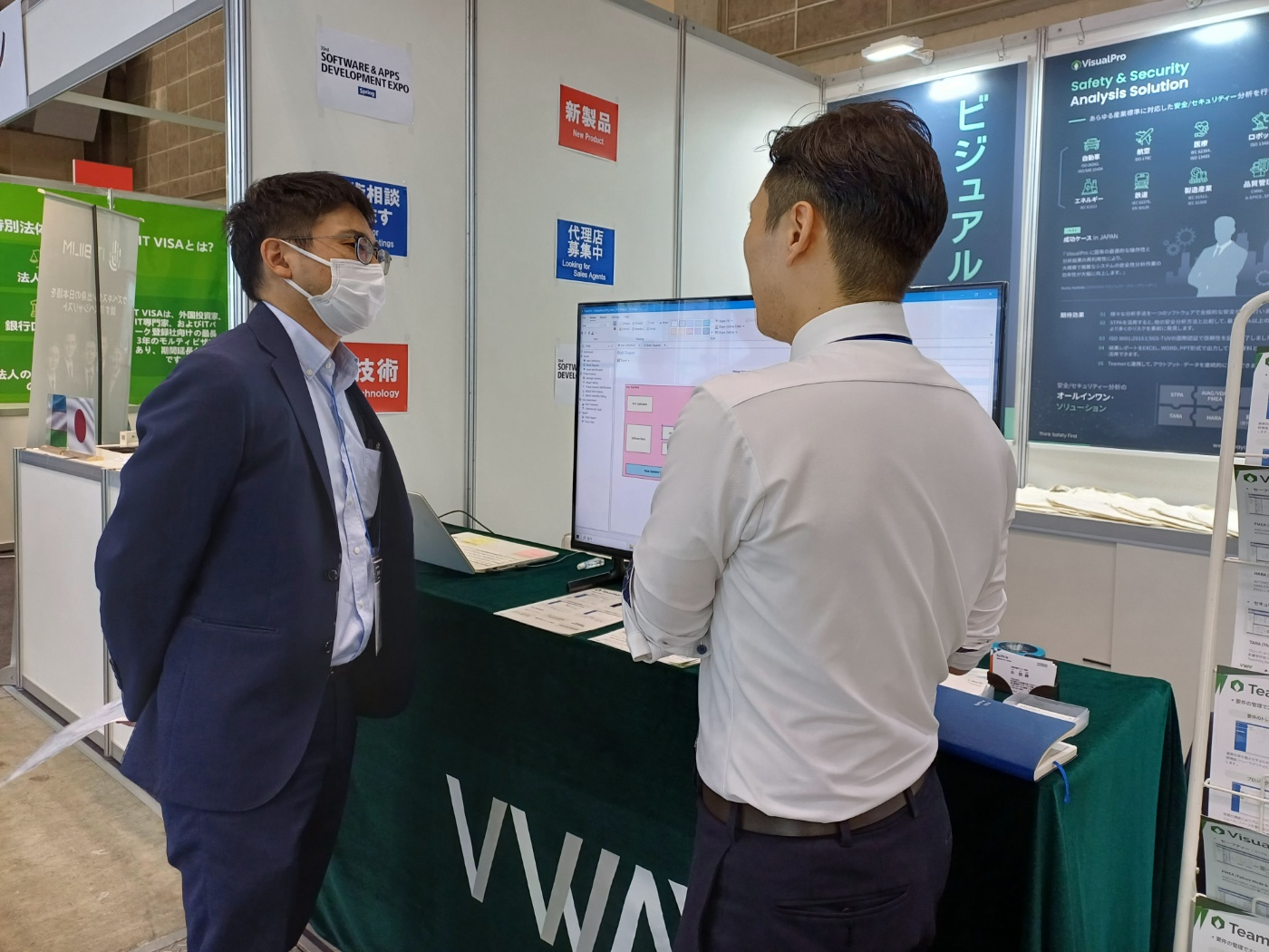 Japan IT Week 2024 Spring 브이웨이 부스 상담 장면 — 모니터에 VisualPro 분석 화면, 뒤편에 일본어 VisualPro 안전·보안 분석 솔루션 패널
