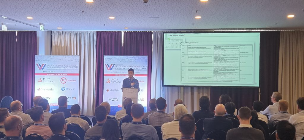독일 베를린 ISO 26262 & SOTIF Conference 2023 발표 장면 — 'STPA on VCU System'의 VCU System Losses 표를 화면에 띄우고 발표하는 모습. 무대 옆 후원사 배너에 Jama Software·UL Solutions·MathWorks·acue·VWAY·Arteris IP